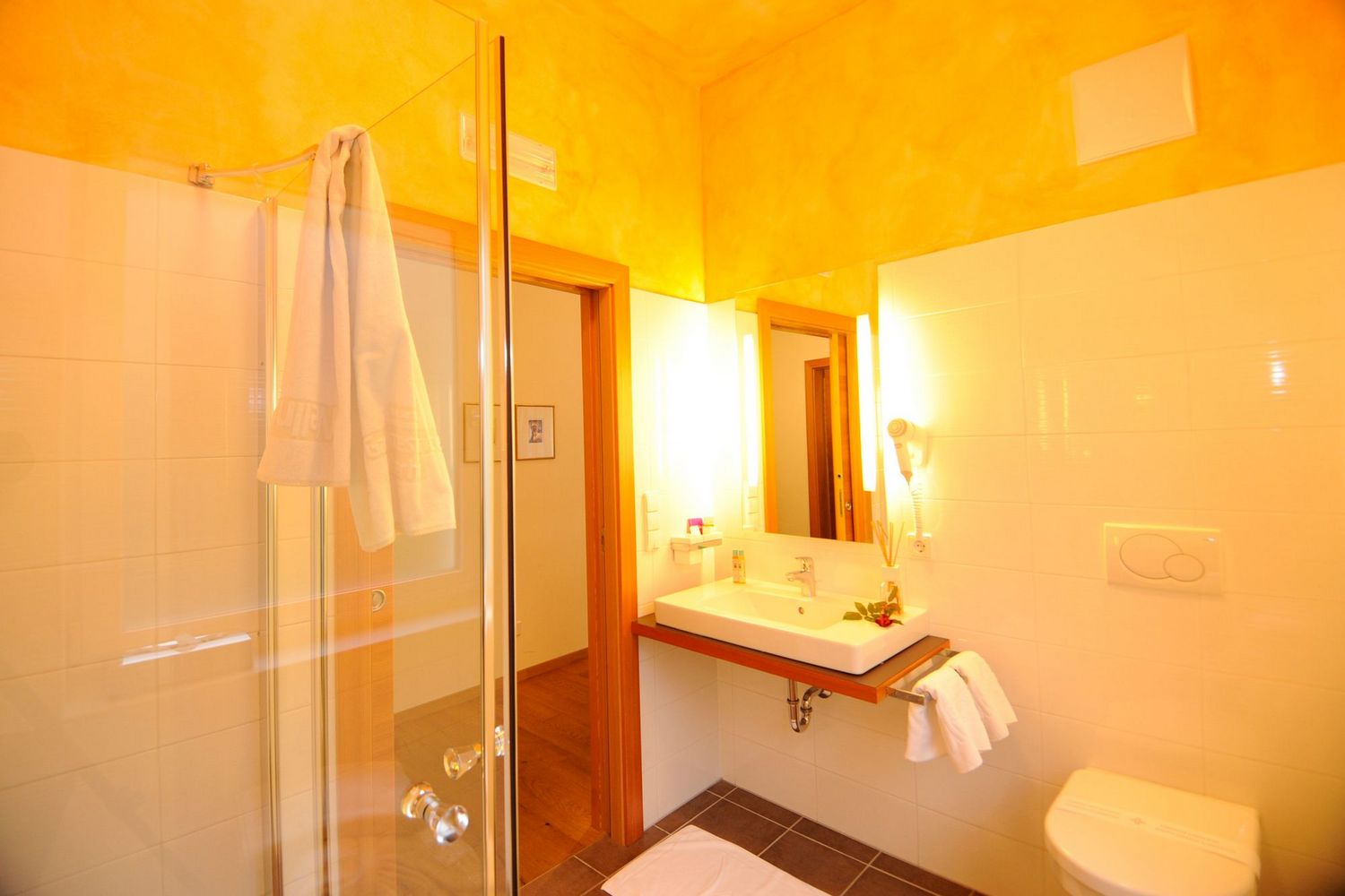 Blick in ein Badezimmer mit einer Glasdusche links, einem Waschbecken mit Spiegel an der Wand und einer Toilette rechts, alles in warmen Gelbtönen gehalten.
