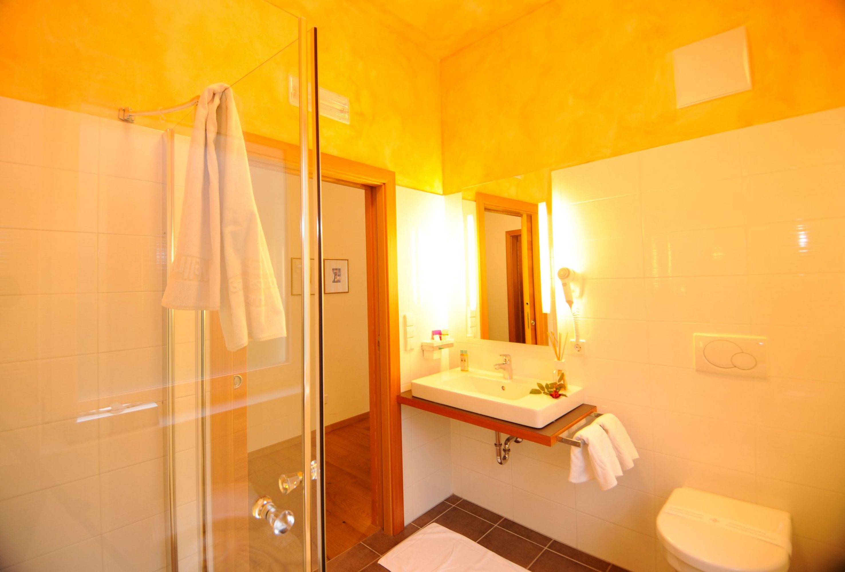 Blick in ein Badezimmer mit einer Glasdusche links, einem Waschbecken mit Spiegel an der Wand und einer Toilette rechts, alles in warmen Gelbtönen gehalten.