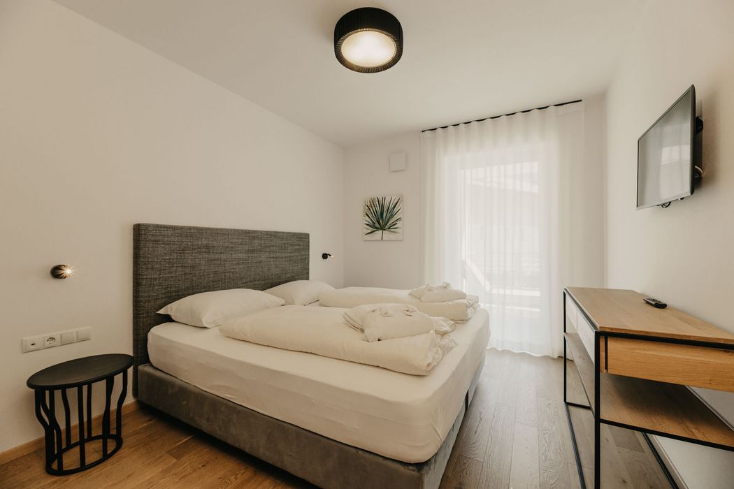Ein minimalistisch eingerichtetes Hotelzimmer mit einem Doppelbett, einem kleinen schwarzen Beistelltisch, einem Fernseher an der Wand und bodentiefen Vorhängen.