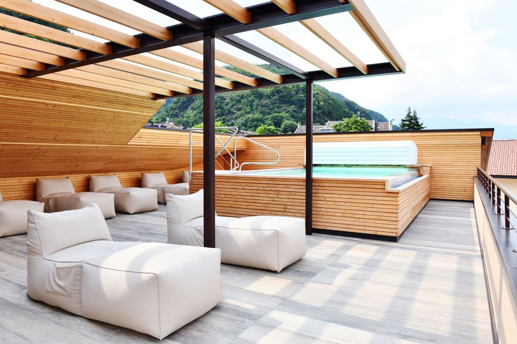 Ampia terrazza sul tetto con rivestimento in legno e pergola, dotata di diversi comodi pouf bianchi e una piscina interrata con bordo in legno, con colline boschive sullo sfondo.