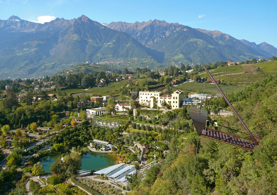 Ein Blick auf Schloss Trauttmansdorff und die umliegenden botanischen Gärten bei Meran, mit einer freischwebenden Aussichtsplattform im Vordergrund und hohen Bergen im Hintergrund.