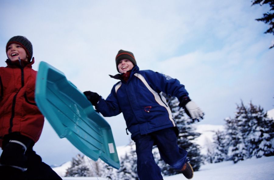 Zwei Kinder in Winterkleidung laufen mit einem blauen Plastikschlitten durch eine verschneite Landschaft.