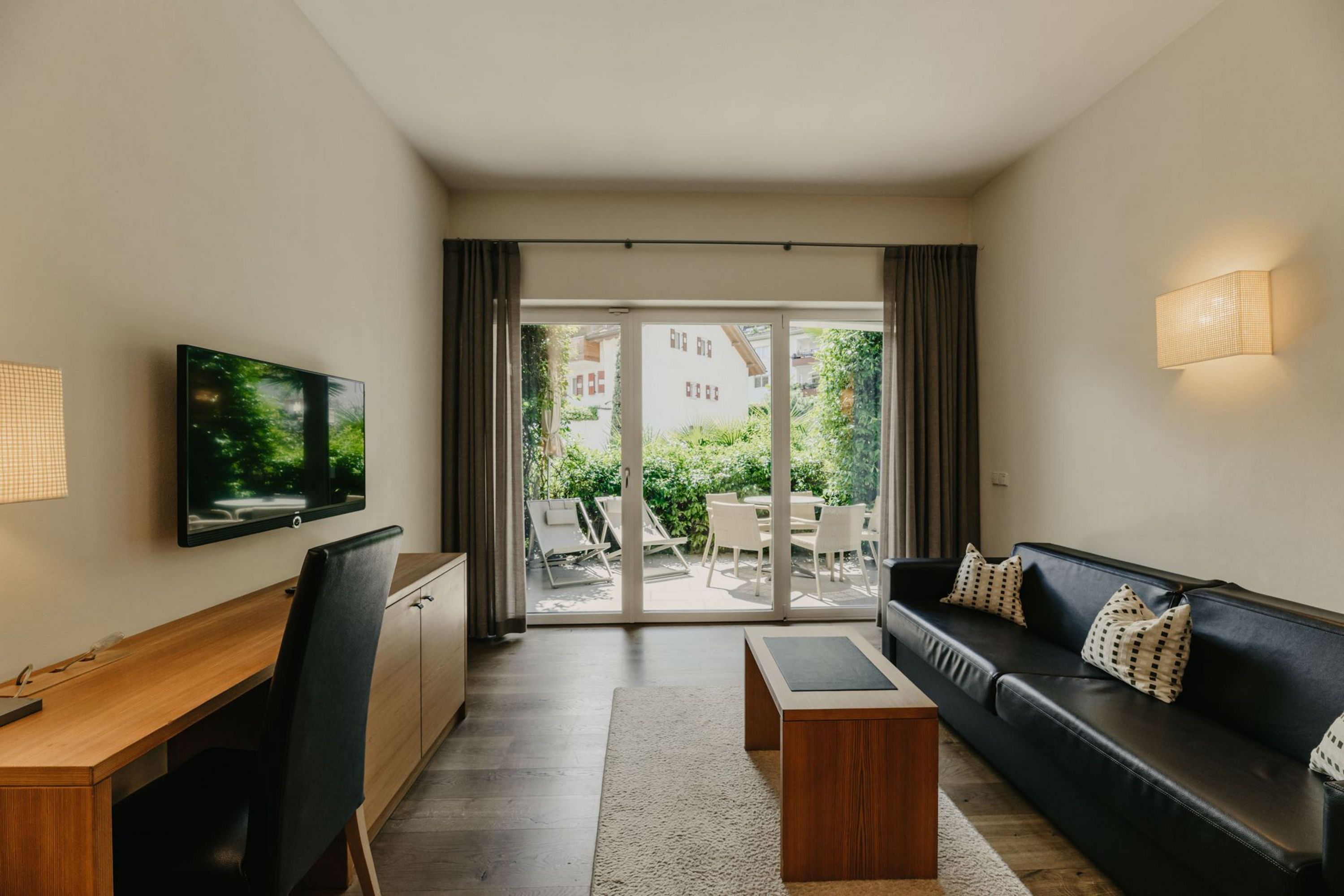 Ein modern eingerichtetes Hotelzimmer mit Schreibtisch, schwarzem Sofa, Couchtisch und einer großen Fensterfront, die auf eine sonnige Terrasse blickt.