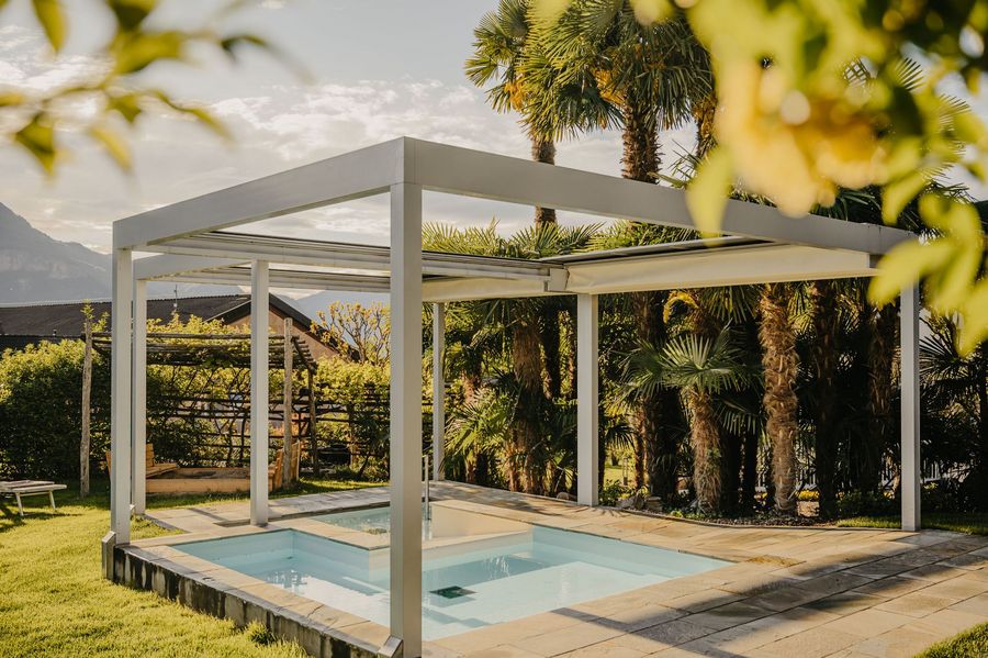 Ein Außenpool mit weißem Pergola-Rahmen in einem sonnigen Garten mit Palmen im Hintergrund.