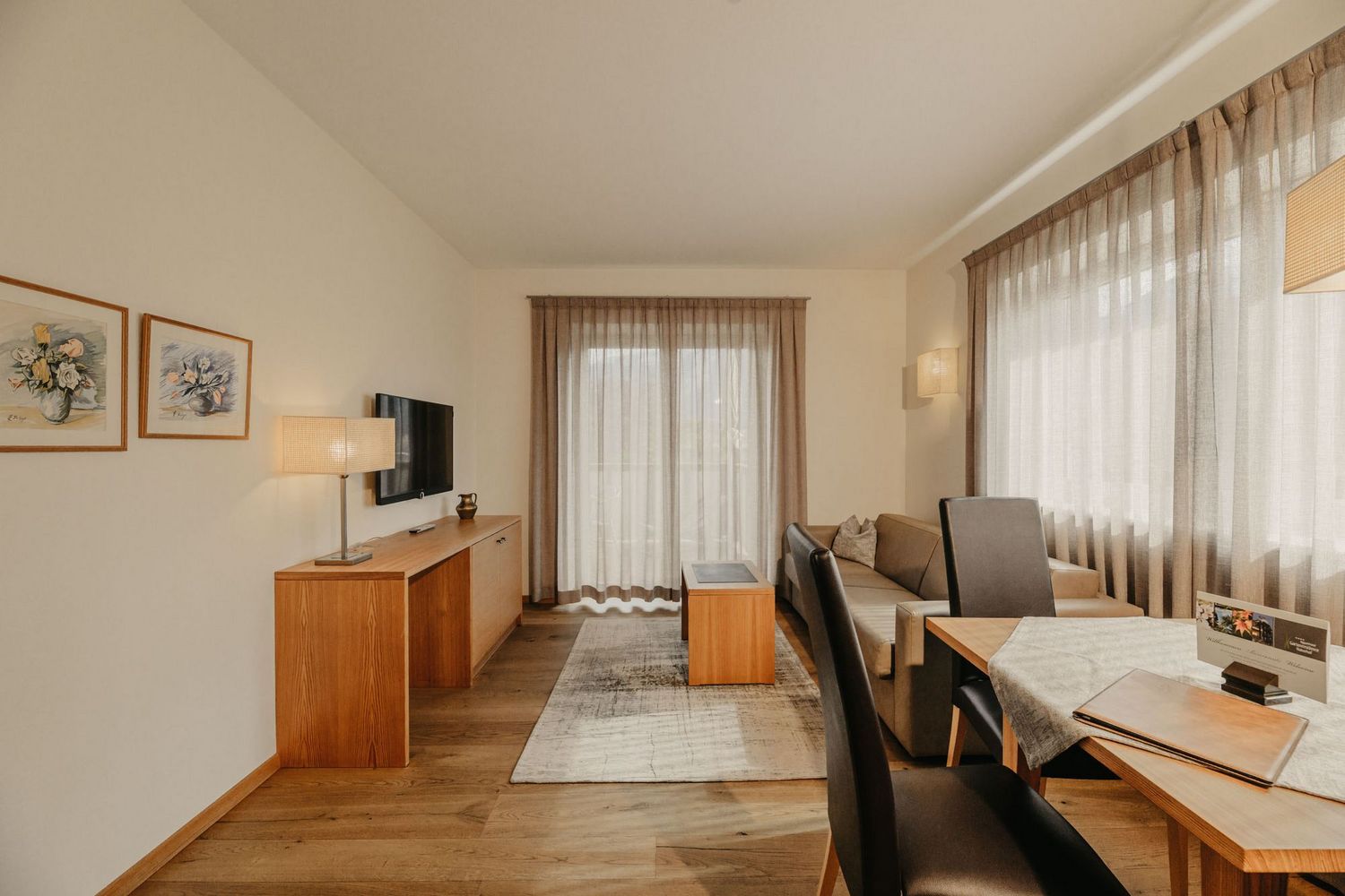 Ein helles Hotelzimmer mit einer Sitzecke bestehend aus einer beigen Couch, einem Holztisch, einem angrenzenden Essbereich mit schwarzen Stühlen und einem Sideboard mit Fernseher.