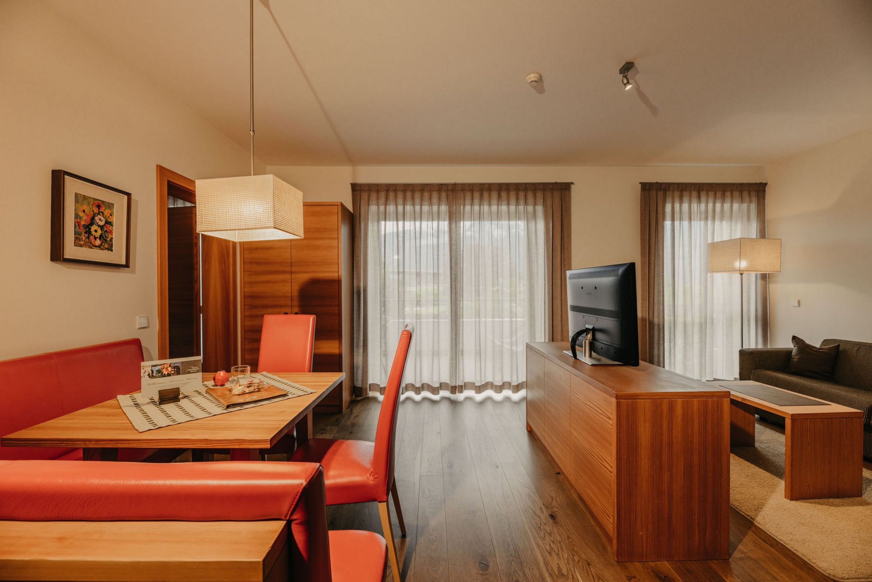 Ein geräumiges Wohn-Esszimmer mit einer roten Eckbank, einem Holzesstisch, einem Fernseher auf einem Sideboard und einer gemütlichen Sitzgruppe bei durchscheinenden Vorhängen.