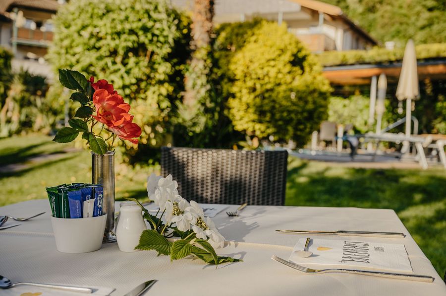 Ein weiß gedeckter Tisch im Freien mit roten Rosen, weißen Blüten, Besteck und Zuckerpäckchen vor einer grünen Gartenkulisse.