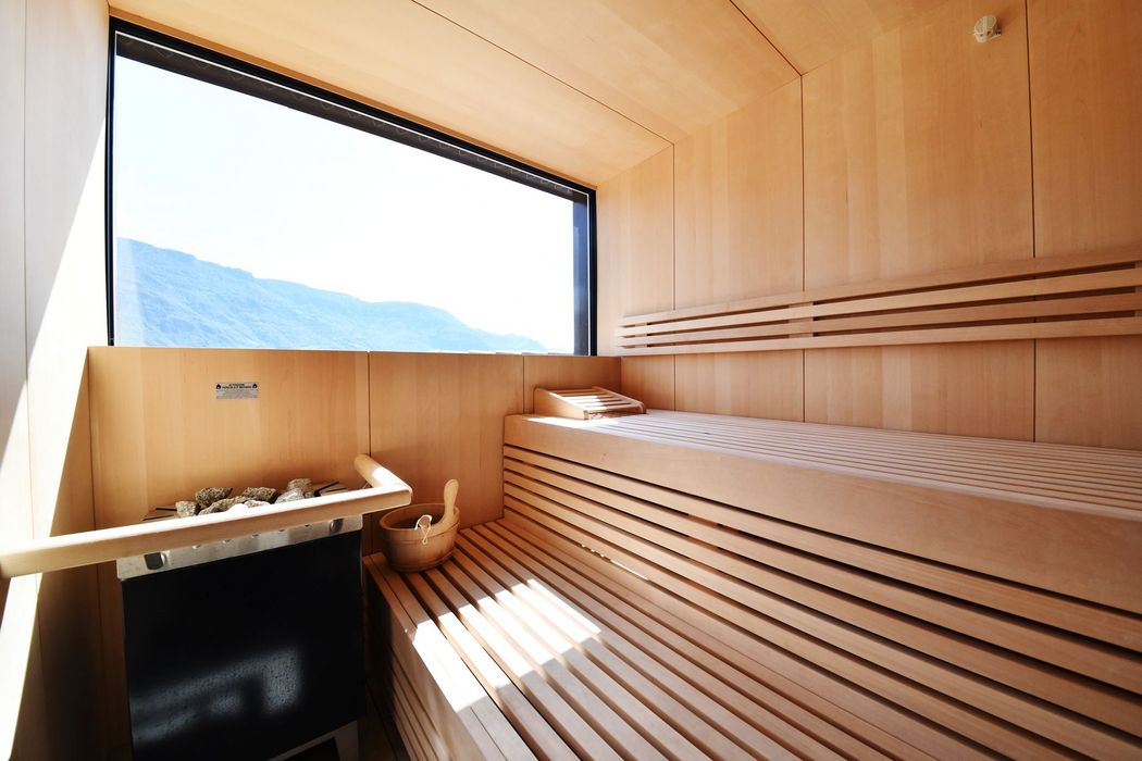 Vista interna di una sauna moderna e luminosa in legno con stufa, secchio in legno e una grande finestra con vista panoramica sulle montagne.