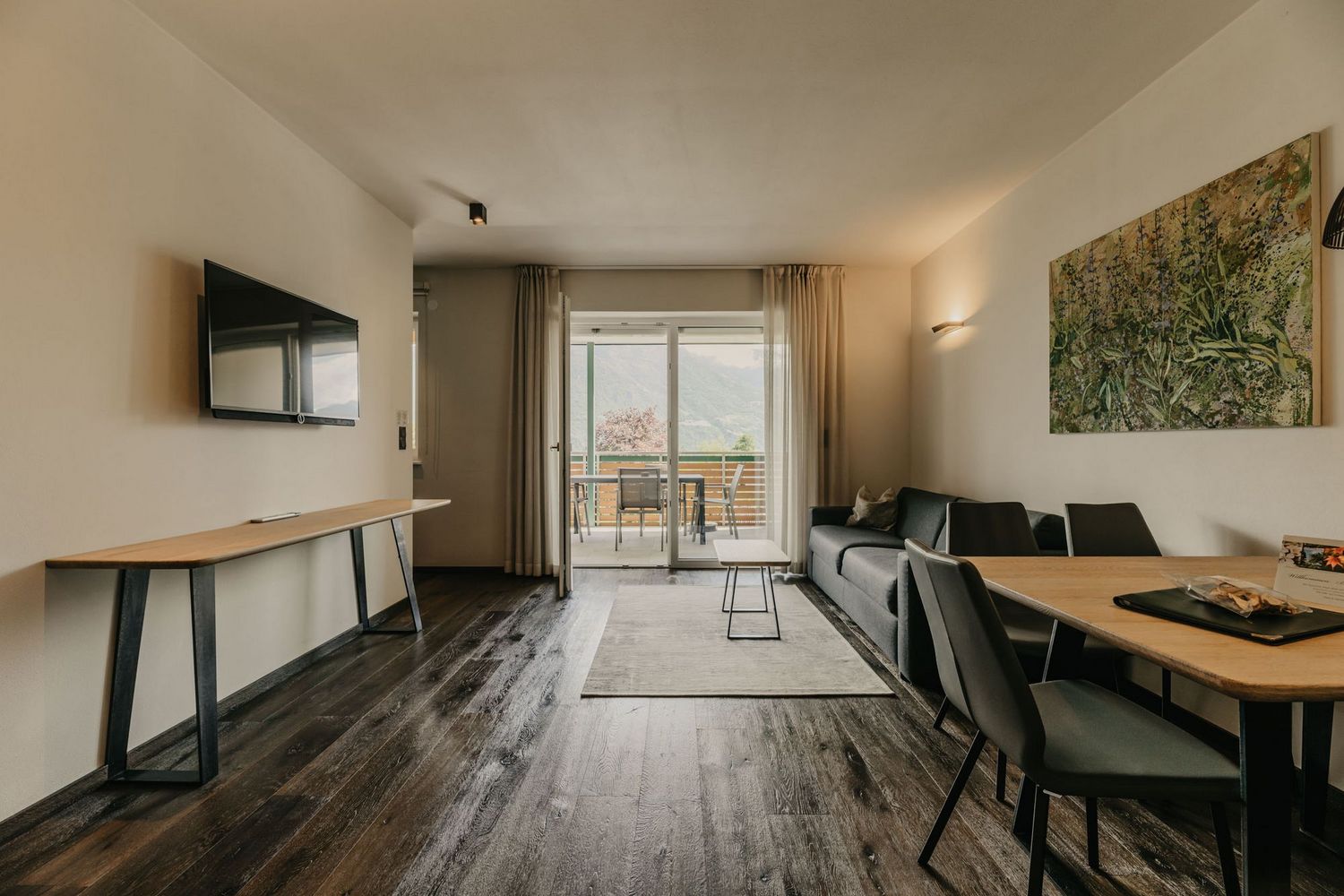 Ein modern eingerichtetes Wohnzimmer mit einem Sofa, einem Esstisch und großen Fenstertüren, die auf einen Balkon mit Bergblick führen.