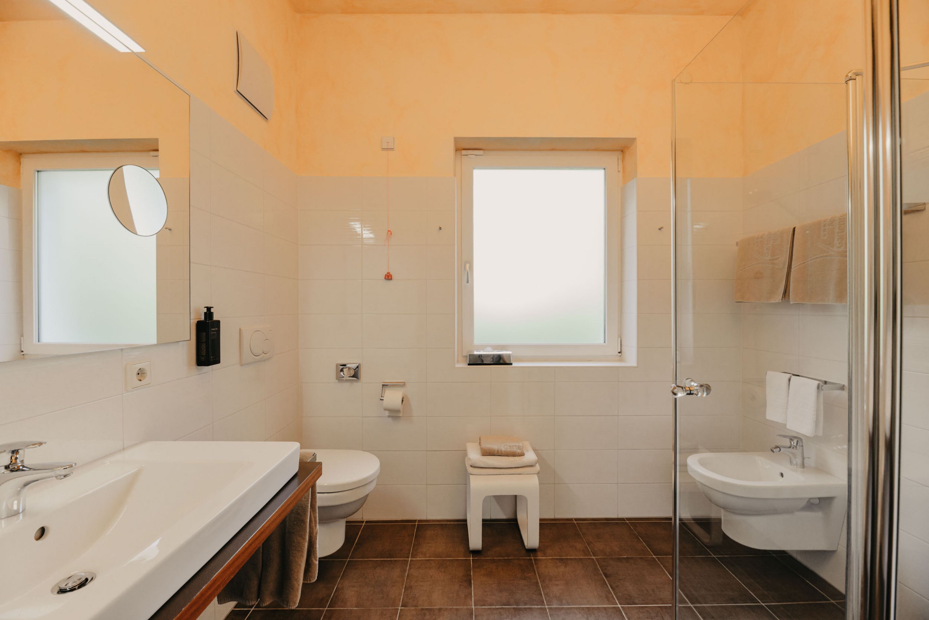 Ein helles Badezimmer mit weiß gefliesten Wänden, beigen Farbakzenten, Waschbecken, Toilette, Bidet und offener Glasdusche.