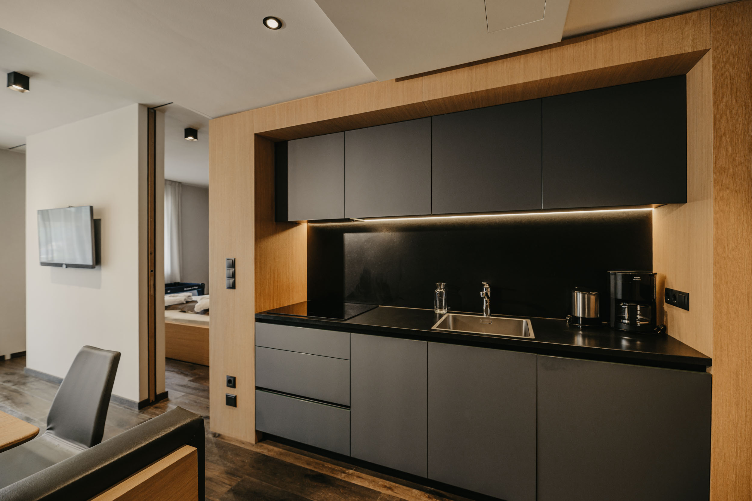 Eine moderne Küchenzeile mit mattschwarzen Schränken, einer dunklen Arbeitsplatte und hellem Holzrahmen, daneben ein Durchgang zu einem Hotelzimmer.
