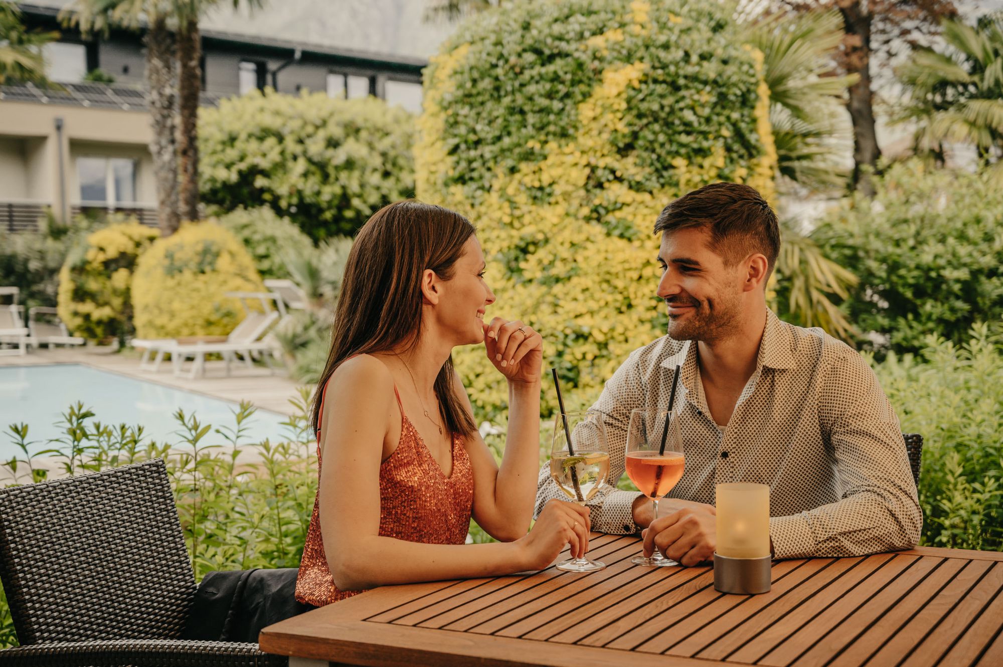 Ein junges Paar sitzt an einem Holztisch im Freien, hält Cocktails in der Hand und schaut sich lächelnd an, im Hintergrund ein Pool und Garten.