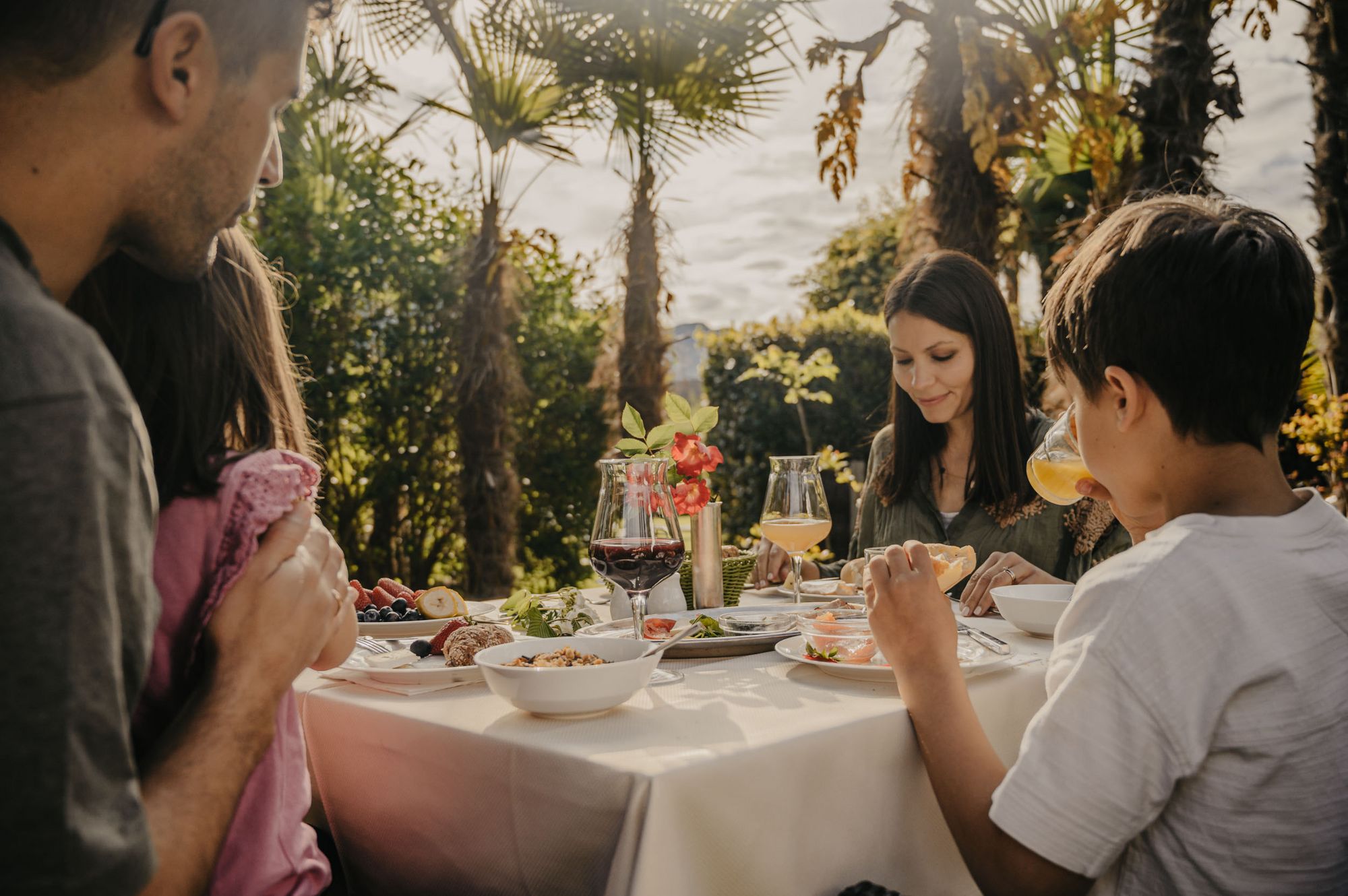 Eine vierköpfige Familie sitzt gemeinsam an einem gedeckten Tisch im Freien unter Palmen und isst bei sonnigem Wetter.