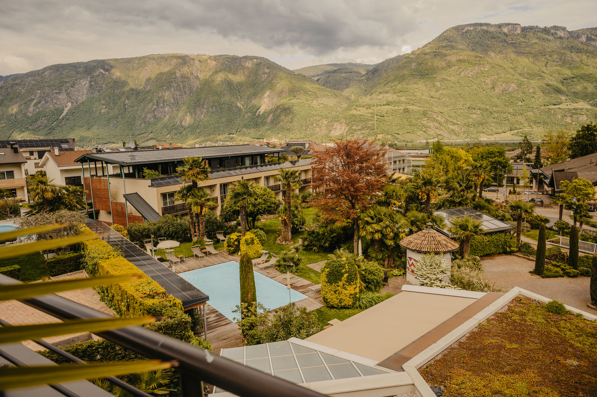 Ein von einem Balkon aus fotografierter Außenbereich einer Hotelanlage mit einem rechteckigen Pool, vielen Palmen und Gartenanlagen, vor einem großen Bergpanorama.