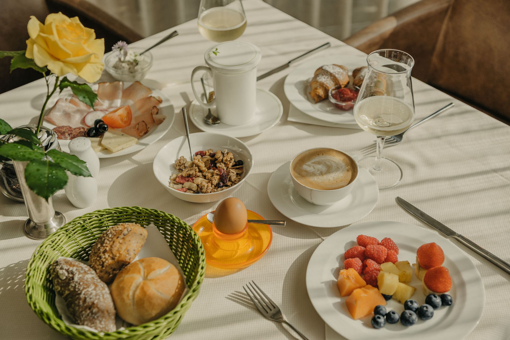 Una tavola riccamente imbandita per la colazione con panini nel cestino, muesli, un uovo sodo, frutta assortita, affettati, formaggi, croissant, caffè e un bicchiere di spumante.