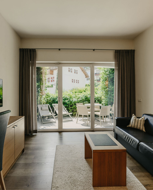 Un soggiorno minimalista con divano in pelle scura, tavolo in legno e vetrate a tutta altezza che conducono a una terrazza soleggiata con mobili da giardino.