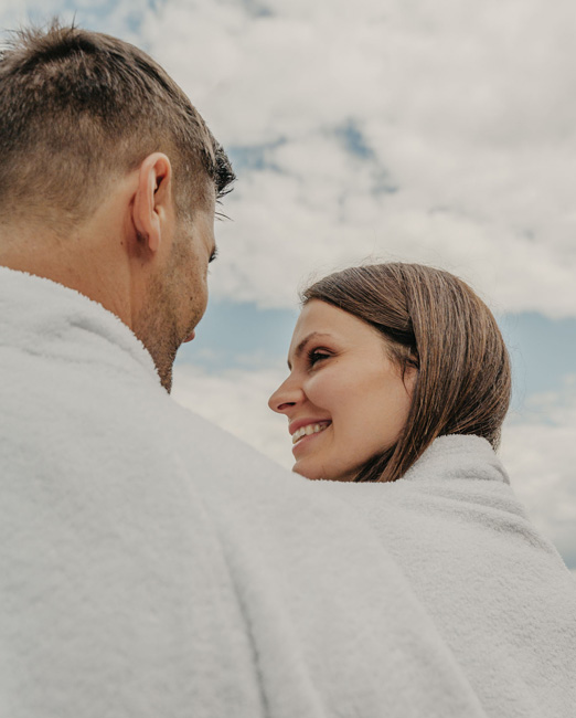 Un uomo e una donna con accappatoi chiari si guardano sorridenti davanti a un cielo nuvoloso.